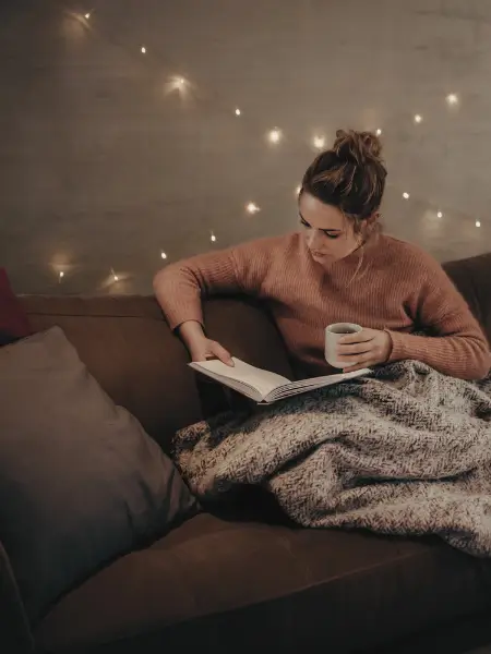 Woman reading on a sofa with a mug and blanket, warm fairy lights in the background.