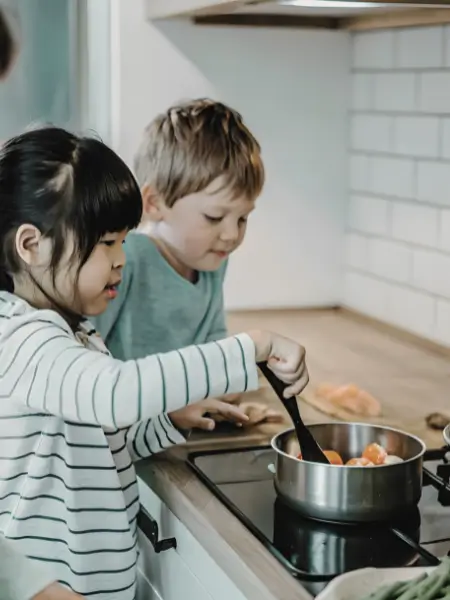 Two children cooking together at a kitchen hob, stirring vegetables in a saucepan.