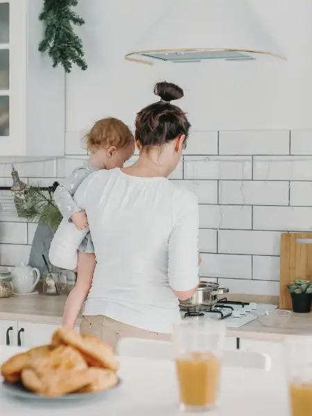 Mother holding a toddler while cooking at the stove in a bright white kitchen.