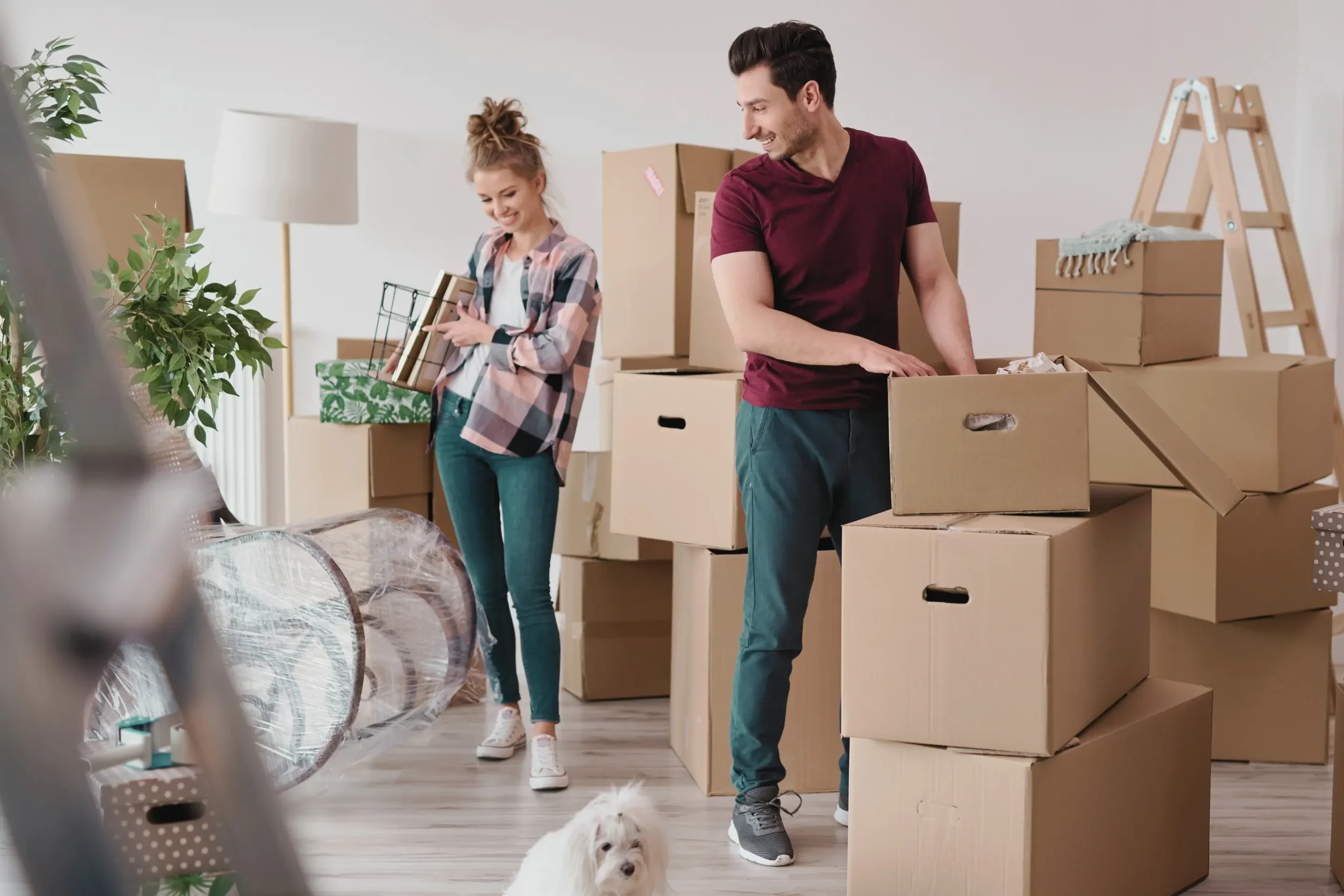 Couple unpacking boxes in a new home, surrounded by moving supplies, with a white dog sitting in the foreground.