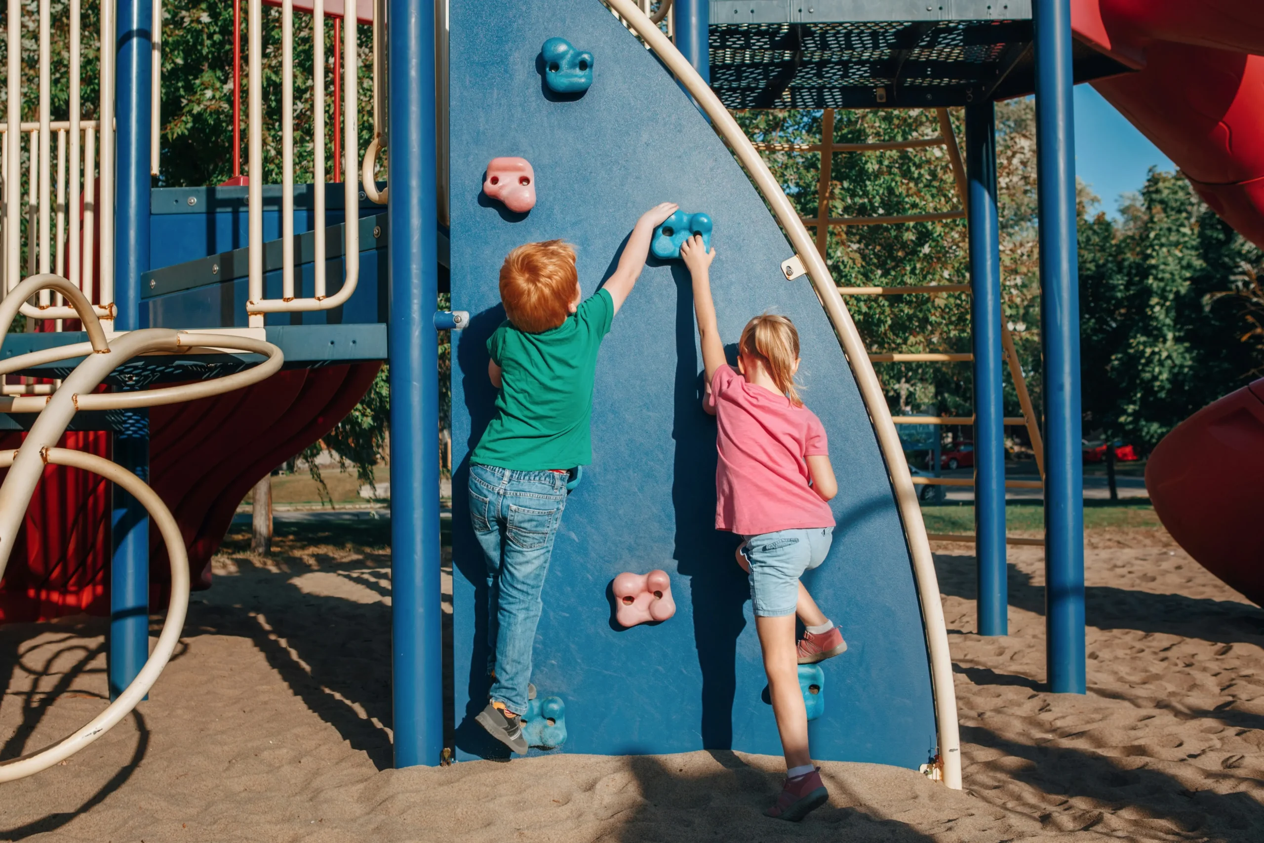 Two children climbing a blue play wall at a playground with sand and colourful climbing structures.