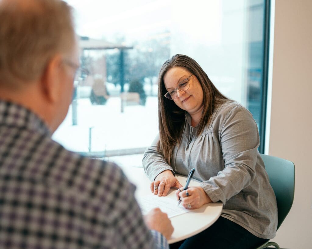 Woman sitting at a table signing paperwork while speaking with a man across from her in a bright office.