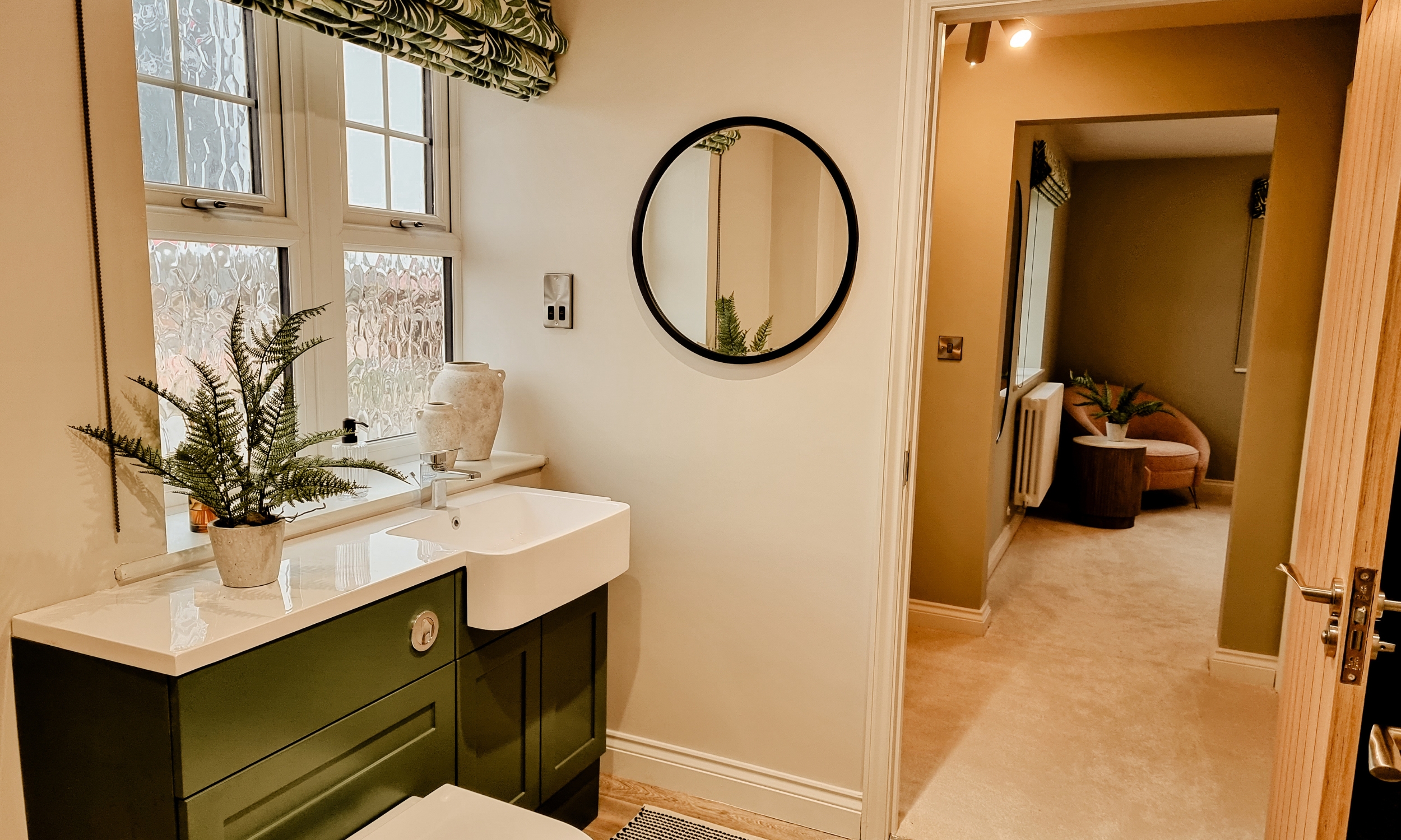 A stylish bathroom featuring green cabinetry, a round mirror, and leafy window blinds, creating a calm and refreshing space in a Broadgate home at The Furlongs.