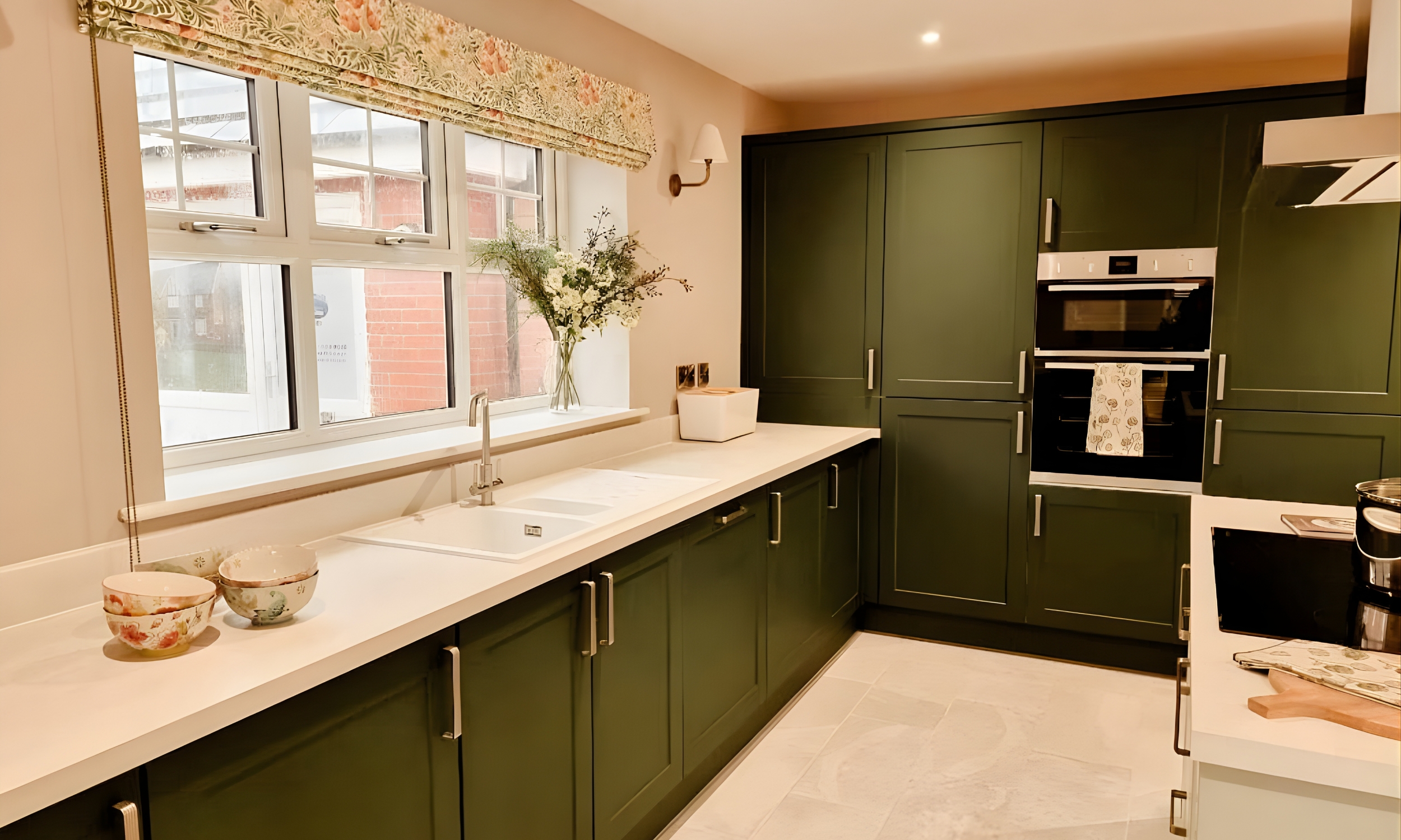 Modern kitchen with deep green cabinetry, white worktops, and floral blinds - reflecting Broadgate’s eye for craftsmanship and calm design.