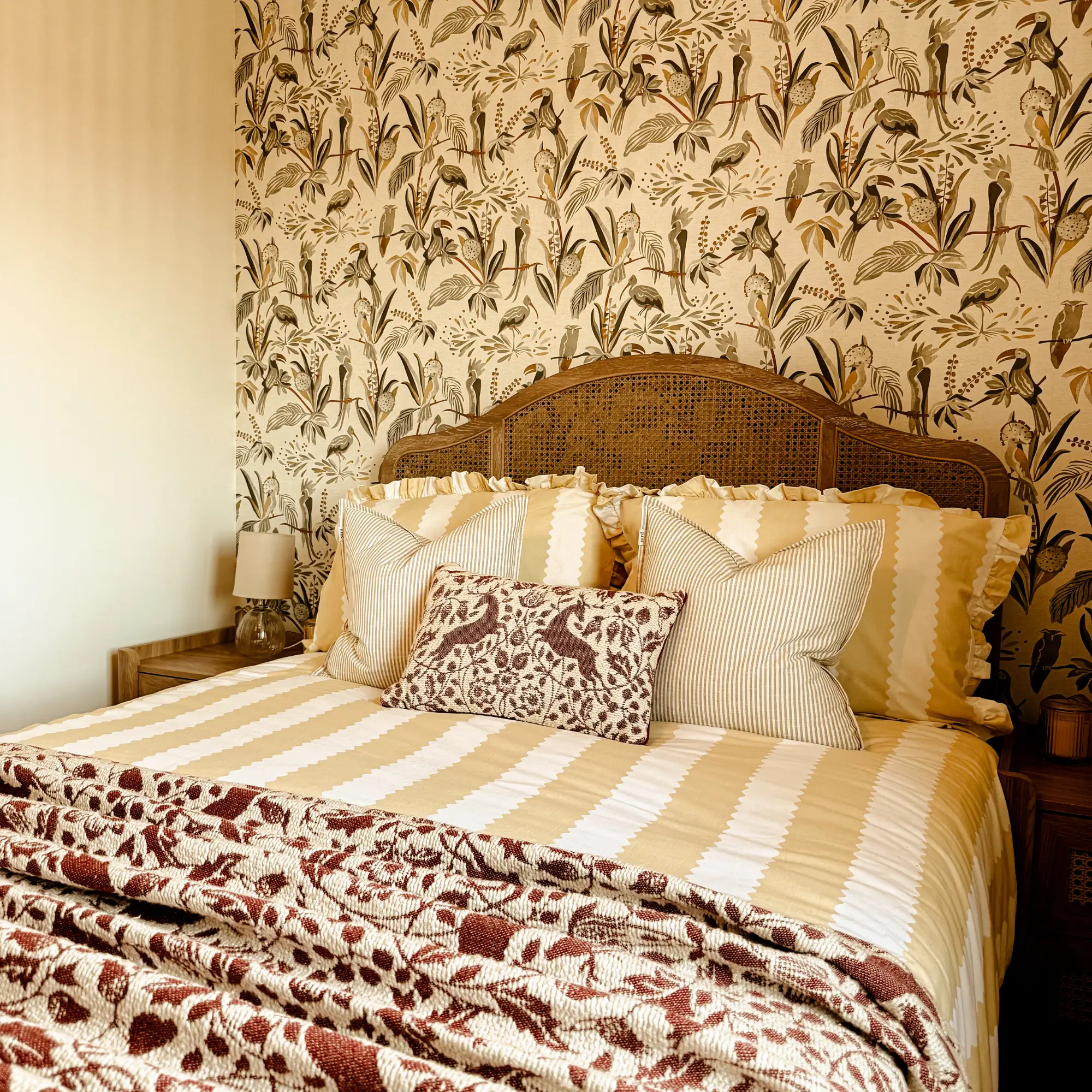 Cosy bedroom with rattan headboard, botanical wallpaper and layered soft furnishings in warm, earthy tones.