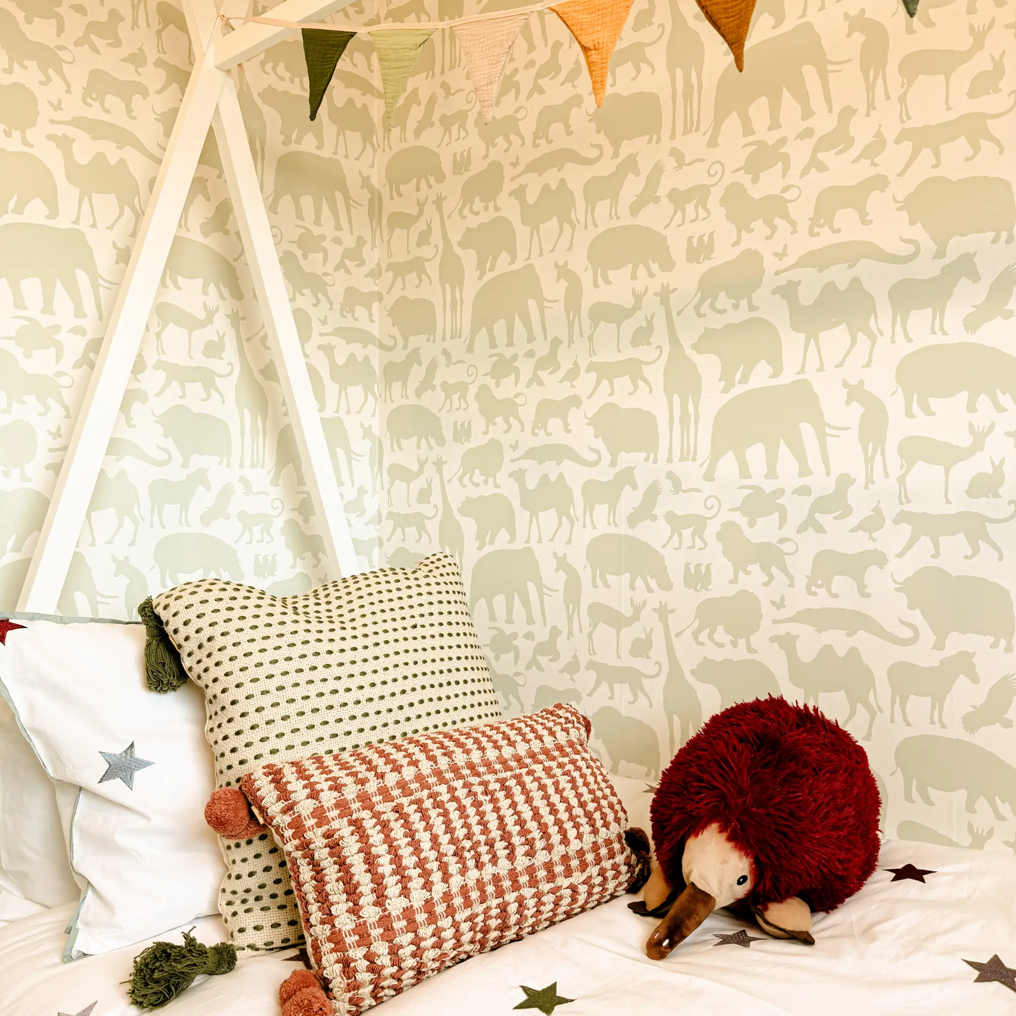 Children’s bedroom corner featuring animal-patterned wallpaper, bunting, playful cushions and a soft toy platypus on the bed.