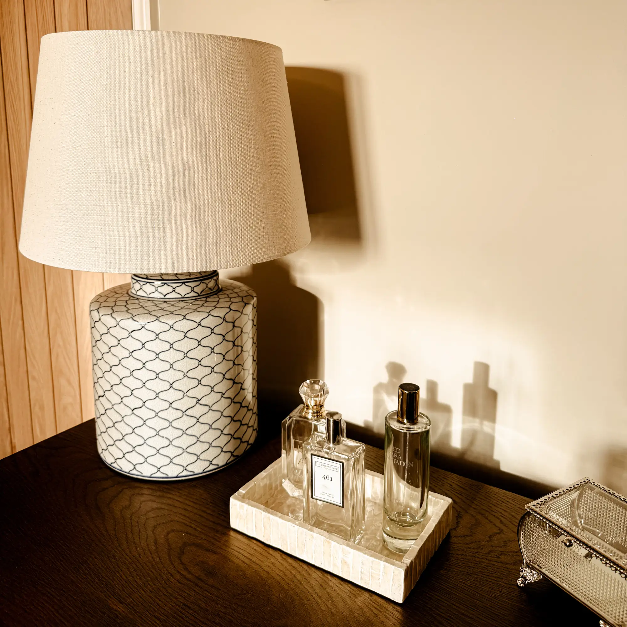 Tabletop vignette showing a ceramic lamp with a neutral fabric shade beside a small tray holding perfume bottles, styled on a dark wooden surface in soft natural light.