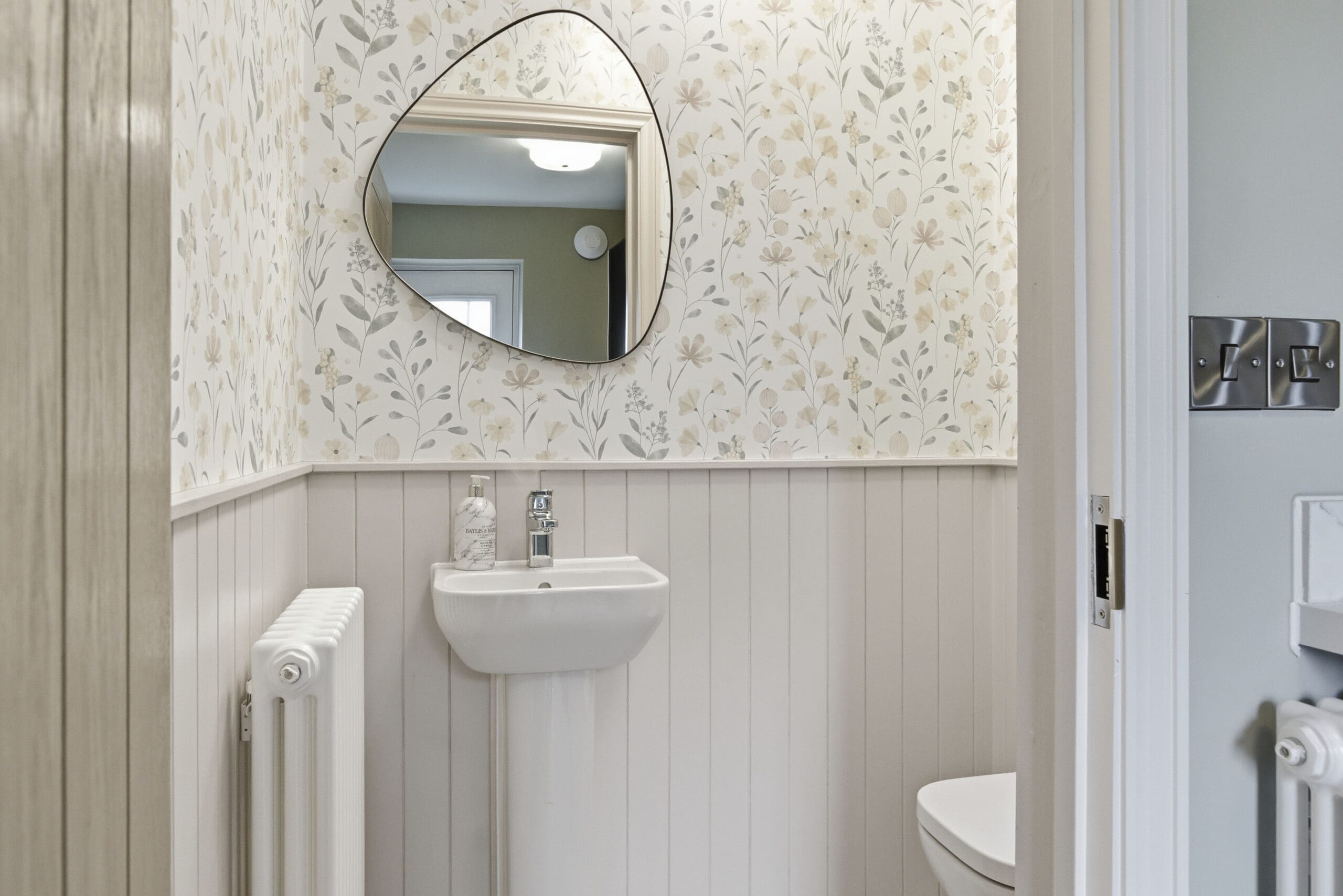 Stylish cloakroom with floral wallpaper, wall panelling, compact basin, and oval mirror creating an elegant guest bathroom space.