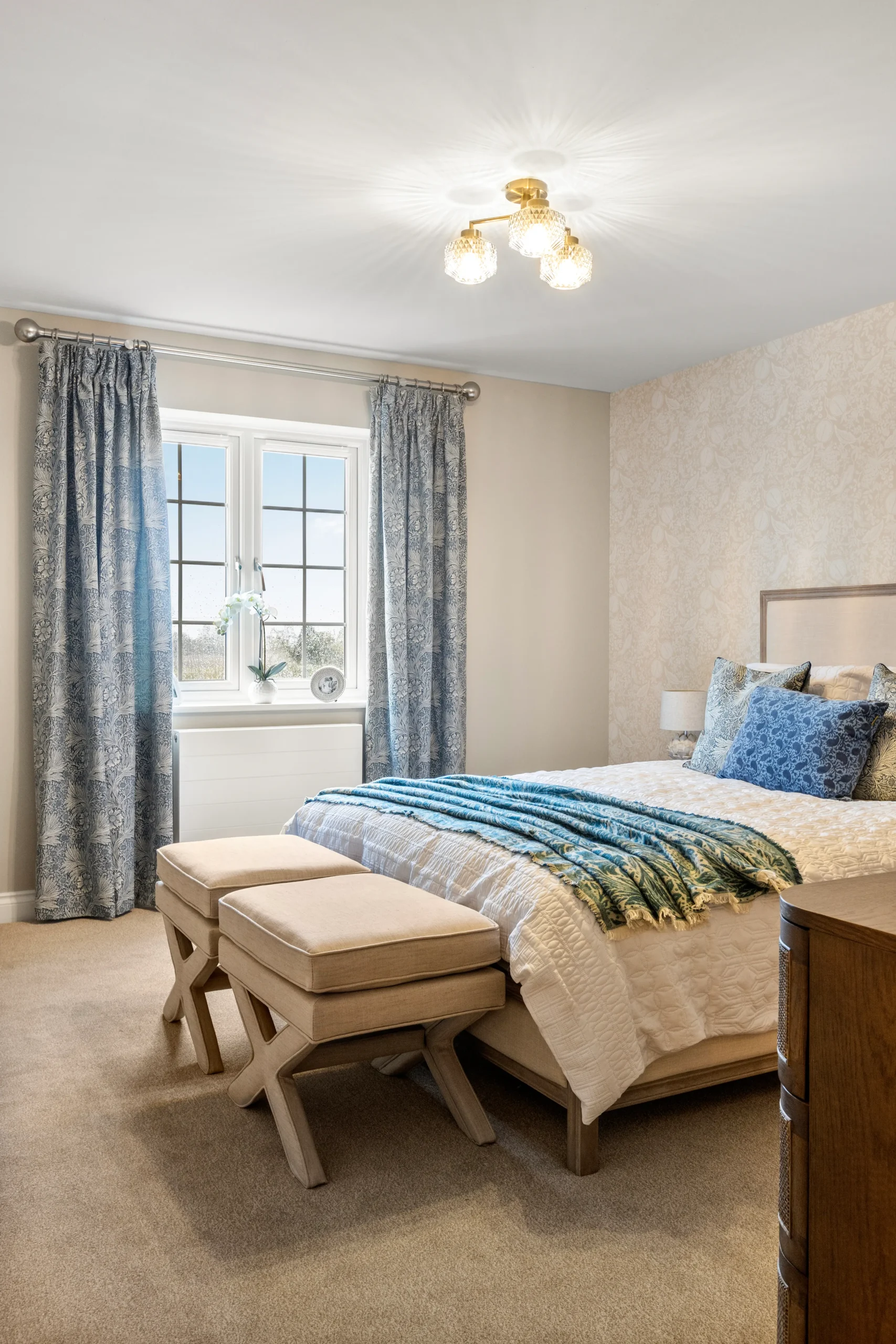 Master bedroom at The Furlongs, Holland Park, Spalding, with William Morris-style blue floral curtains, upholstered headboard, and brass ceiling light fitting.