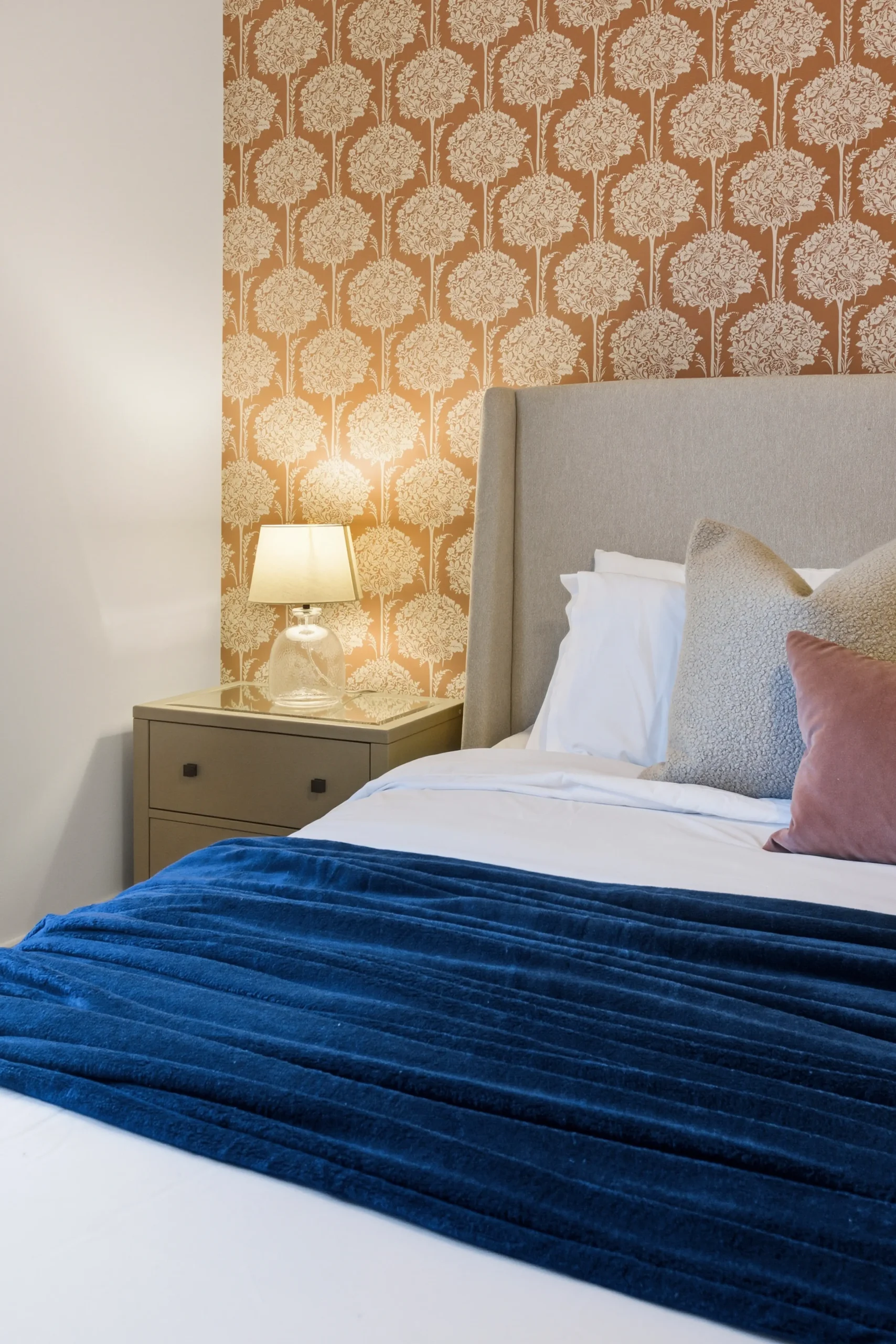 Master bedroom detail at Gleneagles Drive, Woodhall Spa, with terracotta floral feature wallpaper, upholstered headboard, and bedside lamp.
